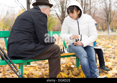 Due amici a giocare a scacchi nel parco con un uomo anziano in un soprabito e più giovane donna seduta su una panchina con la scacchiera tra loro. Foto Stock