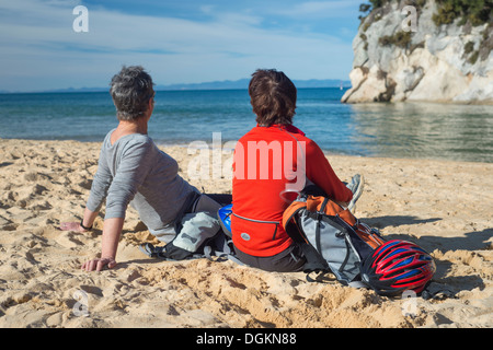 Due donne (età 50+) sulla spiaggia in Nuova Zelanda chiacchierando e guardando il mare, con ciclo caschi e indossando indumenti attivo Foto Stock