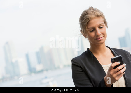 Una bionda e giovane imprenditrice su una città di New York Street. Indossa una giacca nera. Utilizzando un telefono intelligente. Foto Stock