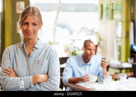 Un giovane in una città coffee shop. Una donna seduta la verifica di un telefono intelligente. Un uomo in piedi con le braccia incrociate. Foto Stock