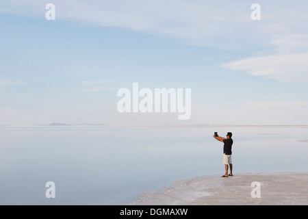 Un uomo in piedi sul bordo dell'allagato Bonneville Saline al tramonto, scattare una fotografia con un dispositivo tablet, vicino Wendover. Foto Stock