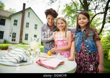 Un estate incontro di famiglia in una fattoria. Una donna e due bambini in piedi fuori da una tabella, che stabilisce la tabella. Facendo una limonata. Foto Stock