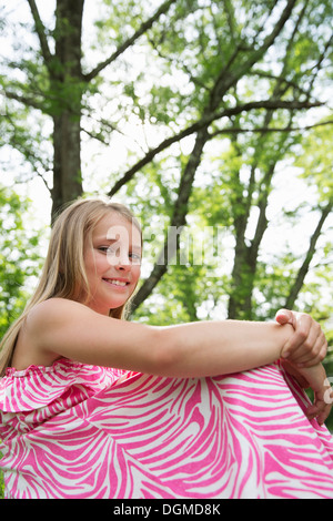 Una giovane ragazza in un rosa sundress modellato seduta sul prato sotto gli alberi in un casale giardino. Foto Stock