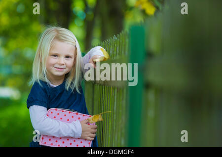 Ragazza 4 anni, tenendo una valigia appoggiata contro una recinzione di legno Foto Stock