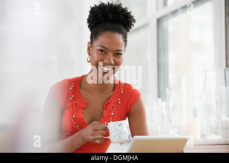 Una giovane donna in maglia rosa utilizzando una tavoletta digitale, tenendo una tazza di caffè. Foto Stock