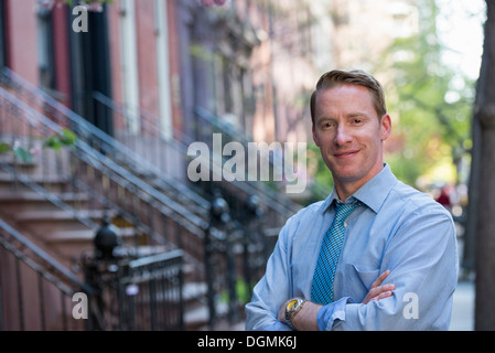 Un Uomo in camicia blu e cravatta blu con le braccia incrociate, in piedi sul marciapiede Foto Stock