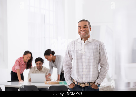 Business. Un uomo in piedi con le mani in tasca e tre persone piegandovi sopra una scrivania dietro di lui. Foto Stock