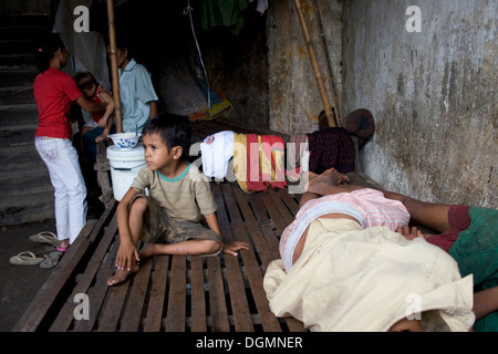 Un giovane bambino operaio ragazzo che vivono in condizioni di povertà è seduta su di un letto in legno mentre gli altri dormono in Kampong Cham, Cambogia. Foto Stock