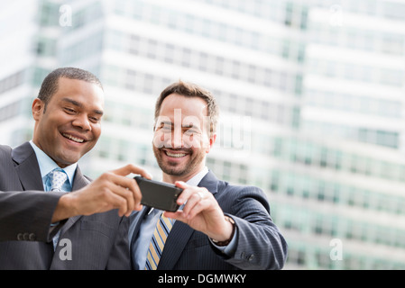 Città. Due uomini in abiti aziendali, guardando uno smart phone, sorridente. Foto Stock