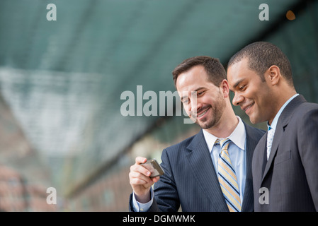 Città. Due uomini in abiti aziendali, guardando uno smart phone, sorridente. Foto Stock