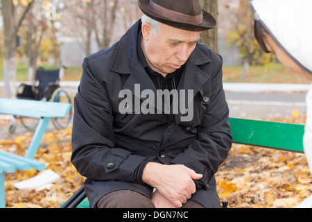 Premuto lonely vecchio seduto nel suo sovrarivestimento su una panchina nel parco con le mani giunte fissando morosely a terra nel profondo del pensiero. Foto Stock