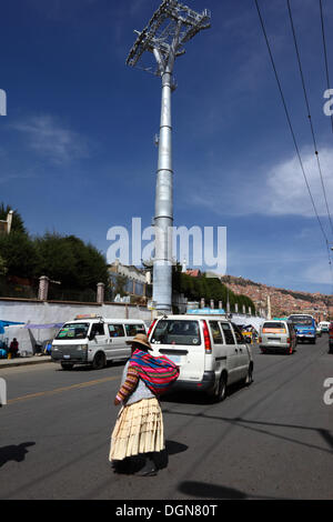 LA PAZ, Bolivia, 23 Ott, 2013. Una signora Aymara attraversa la strada di fronte a uno dei piloni accanto al cimitero principale per la nuova cabinovia / telecabina attualmente in costruzione per il collegamento delle città di La Paz e El Alto. Il sistema è stato costruito dalla società austriaca Doppelmayr ad un costo di $234.6 milioni di euro finanziati dal governo boliviano. Credito: James Brunker / Alamy Live News Foto Stock