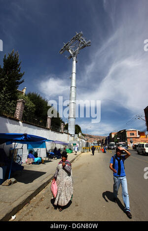 LA PAZ, Bolivia, 23 Ott, 2013. Una signora Aymara cammina per la strada di fronte a uno dei piloni accanto al cimitero principale per la nuova cabinovia / telecabina attualmente in costruzione per il collegamento delle città di La Paz e El Alto. Il sistema è stato costruito dalla società austriaca Doppelmayr ad un costo di $234.6 milioni di euro finanziati dal governo boliviano. Credito: James Brunker / Alamy Live News Foto Stock