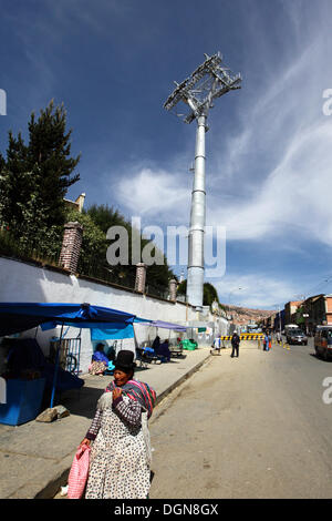 LA PAZ, Bolivia, 23 Ott, 2013. Una signora Aymara cammina per la strada di fronte a uno dei piloni accanto al cimitero principale per la nuova cabinovia / telecabina attualmente in costruzione per il collegamento delle città di La Paz e El Alto. Il sistema è stato costruito dalla società austriaca Doppelmayr ad un costo di $234.6 milioni di euro finanziati dal governo boliviano. Credito: James Brunker / Alamy Live News Foto Stock