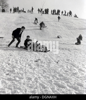 Anni '1960, storia, inverno e gente su una collina che si diverte sulla neve, con una madre e i suoi due figli su una slitta, papà in supporto, Cumbria, Inghilterra, Regno Unito. Foto Stock
