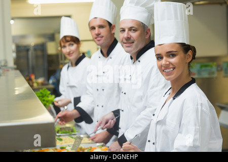 Quattro chef sorridente alla videocamera mentre si lavora al servizio dei vassoi Foto Stock