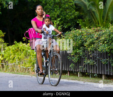 Una donna con un bambino su una bicicletta utilizzando un telefono cellulare, La Digue Island, Seychelles, Africa, Oceano Indiano Foto Stock