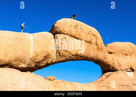 Giovani uomini di arrampicarsi su un arco di roccia, vicino al Spitzkoppe vette di granito, Damaraland, Namibia, Africa Foto Stock