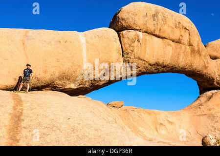 Giovane uomo di arrampicarsi su un arco di roccia, vicino al Spitzkoppe vette di granito, Damaraland, Namibia, Africa Foto Stock