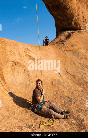 Giovane uomo la discesa in corda doppia da un arco di roccia, appeso a testa in giù, vicino al Spitzkoppe vette di granito, Damaraland, Namibia, Africa Foto Stock