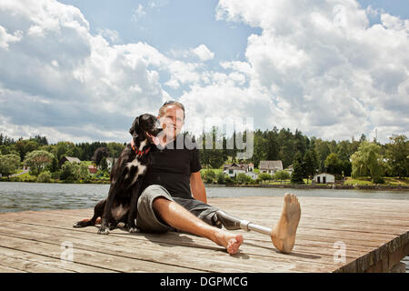 Uomo con una gamba protesico seduto su di un molo con il suo cane da caccia Foto Stock