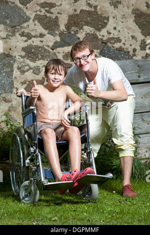 Ragazzo seduto in una sedia a rotelle con il suo amico in piedi accanto a lui Foto Stock