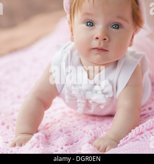 Sweet Little Baby girl giacente sul suo pancino indossa una camicia bianca e berretto lavorato a maglia, guardando la telecamera. Foto Stock