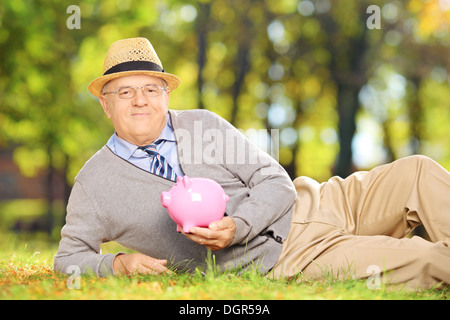 Sorridente gentiluomo maturo giacente su un erba e tenendo un salvadanaio in un parco Foto Stock