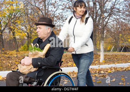 Donna felice aiutare un anziano uomo disabile che lo spinge lungo la strada nella sua sedia a rotelle dopo aver preso lui fuori a fare la spesa. Foto Stock