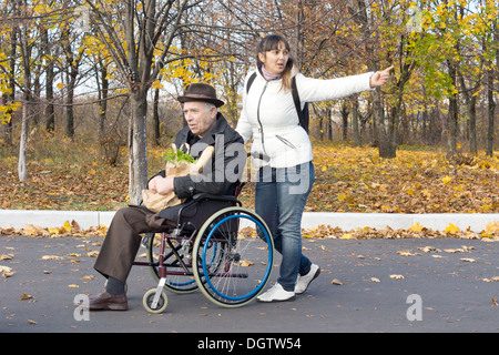 Donna spingendo un uomo anziano in una sedia a rotelle e stirando il suo braccio rivolto a qualcosa e a chiamare in quanto a piedi lungo la strada. Foto Stock