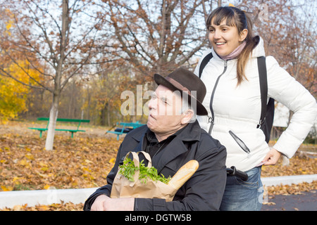 Donna felice con il suo anziano padre disabili sostando in strada per raddrizzare la sua camicia come lei lo spinge lungo nella sua sedia a rotelle mentre sono fuori a fare la spesa. Foto Stock