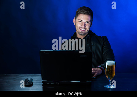 Giovane uomo seduto al buio davanti al suo computer portatile, con una birra accanto a lui, immagine simbolica su computer e dipendenza Foto Stock