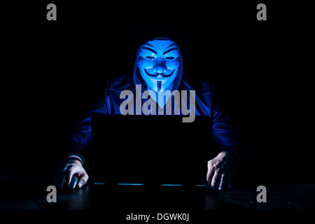 Giovane uomo che indossa una maschera anonimo e una cappa seduto davanti a un computer portatile Foto Stock