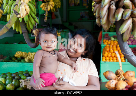 Fornitore femmina mentre tiene il suo bambino, stallo del mercato per la vendita di frutta e verdura, Bentota, Aluthgama, Sri Lanka Foto Stock