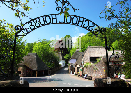 Un villaggio con cottage con il tetto di paglia e di alberi a foglia, incorniciato da un ferro battuto segno 'Scuola casa'. Foto Stock