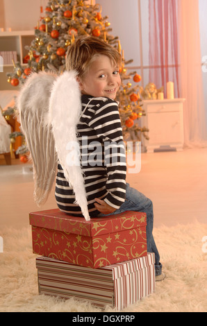 Ragazzo che indossa ali d'Angelo seduto su doni nella parte anteriore di un albero di Natale Foto Stock