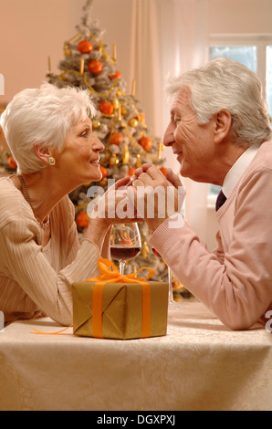 Coppia matura con vino rosso bicchieri e un presente seduta davanti a un albero di Natale Foto Stock