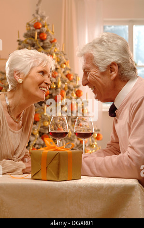 Coppia matura con vino rosso bicchieri e dono seduto davanti a un albero di Natale Foto Stock