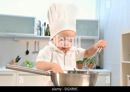 Il Toddler, 2-4, indossando un chef hat in una cucina e tenendo una frusta Foto Stock