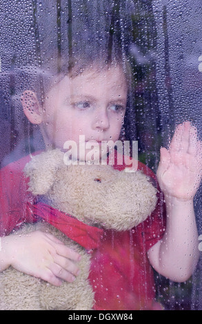 Ragazzo che guarda tristemente fuori di una finestra con gocce d'acqua, tenendo un peluche nelle sue braccia Foto Stock