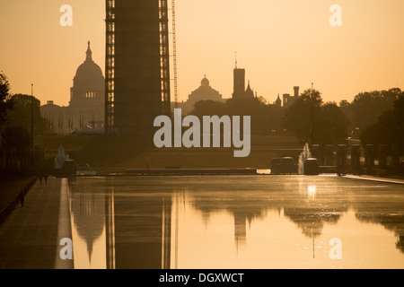 WASHINGTON DC, Stati Uniti - il sole nascente appare basso all'orizzonte dietro il Washington Monument, che è coperto da impalcature a causa di un importante progetto di restauro a seguito di danni sismici. La scena si riflette nelle acque calme della piscina riflettente sul National Mall. Foto Stock