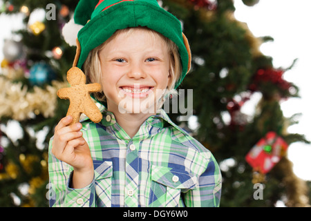 Giovane ragazzo carino sorridente che indossa un elfo cappello di fronte ad un albero di Natale che tiene un uomo di pan di zenzero. Isolato su bianco. Foto Stock