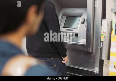 Uomo che utilizza una banca bancomat sulla strada con un ladro a guardare lui da dietro Foto Stock