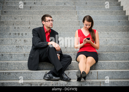 Young business donna seduta sulle scale e lavorare con lo smartphone mentre l uomo è annoiato e arrabbiato Foto Stock