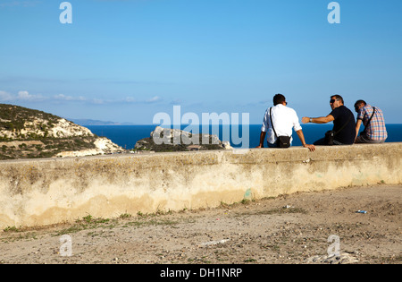 Gli uomini seduti sulla parete dal faro di Capo Sant'Elia a San Bartolomeo guardando sopra Calamosca Bay - Sardegna Foto Stock