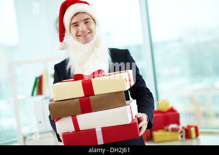 Immagine di happy businessman in Santa Cappello e barba holding pila di doni e guardando la telecamera in ufficio Foto Stock