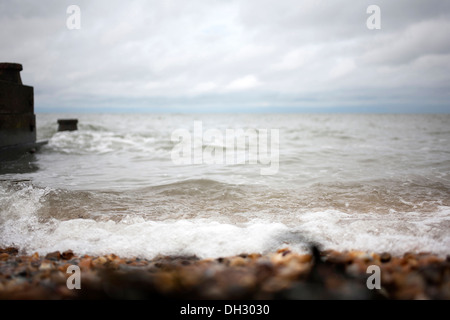 Vista del mare dalla spiaggia di ciottoli su nuvoloso giorno mostrando piccola onda la rottura su pietre e legno scuro groyne posti in mare. Foto Stock