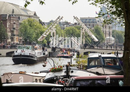 Paesi Bassi, Amsterdam, Barche passando aperta ponte levatoio Skinny Bridge. Di fronte case galleggianti. Torna Carre Theatre, Amstel Hotel Foto Stock