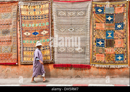 Moquette o tappeti con gli arabi e i simboli di Berbera e modelli, appeso a una facciata per vendita, uomo anziano in una djellabah passato a piedi Foto Stock