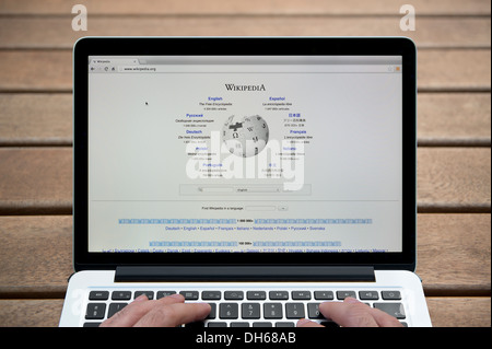 Il sito web di Wikipedia su un MacBook contro una panca in legno sfondo all'aperto tra cui un uomo le dita (solo uso editoriale). Foto Stock